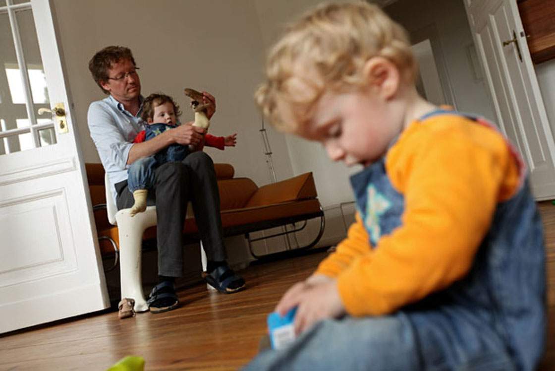 Oliver H., 42, a married federal employee on 6-month paternity leave, puts shoes on his twin 14-month-old daughter Lotte while her sister Alma plays on the floor at his home.