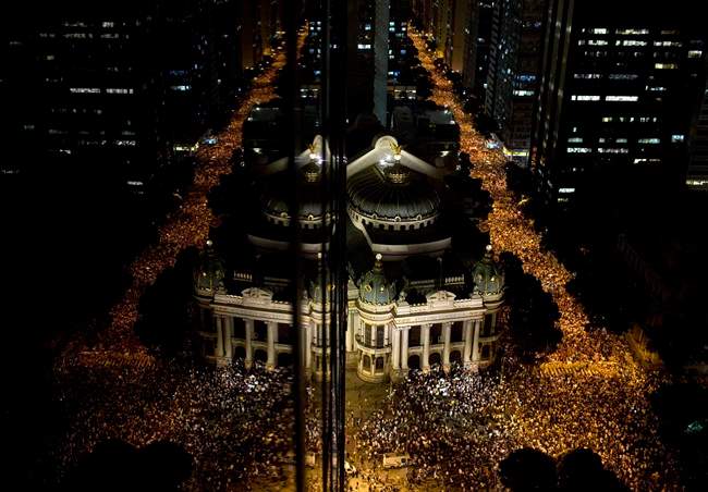 FILE - In this June 17, 2013 file photo, protestors are reflected on the glass of a building, left, for an anti-goverment march during the Confederations Cup soccer tournament in Rio de Janeiro, Brazil. The protests have grown smaller but more violent and show no sign of going away. With financial backing for the 2106 Olympics drying up, Brazilian organizers are scrambling to raise record amounts of local sponsorship money to cover any budget shortfall and avoid a government bailout. Just a few months ago, a top Rio official said $700 million in government aid would be needed to make up for a shortfall in balancing the local operating budget. That’s no longer politically tenable. On-going violent street protests, which began five months ago during the Confederations Cup, the warm-up for next year's World Cup, have focused on Brazil's poor schools, shabby hospitals and soaring costs. Protesters have questioned why the country is spending $15 billion on the World Cup and a similar amount on the Olympics. (AP Photo/Felipe Dana, File)