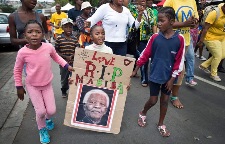 Soweto, Johannesburg, South Africa:A young girl with a placard showing the face of Nelson Mandela and referring to his clan name “Madiba”, marches with others to celebrate his life, in the street outside his old house in Soweto, Johannesburg, South Africa Friday, Dec. 6, 2013. (Photo credit: AP Photo)