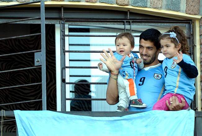 Uruguay's soccer player Luis Suarez, center, with his children Benjamin, left, and Delfina, waves to fans from his home, before the start of his team's World Cup round 16 match with Colombia, on the outskirts of Montevideo, Uruguay, Saturday, June 28, 2014. FIFA banned Suarez from all football activities for four months for biting an opponent at the World Cup, a punishment that rules him out of the rest of the tournament. (AP Photo/Matilde Campodonico)