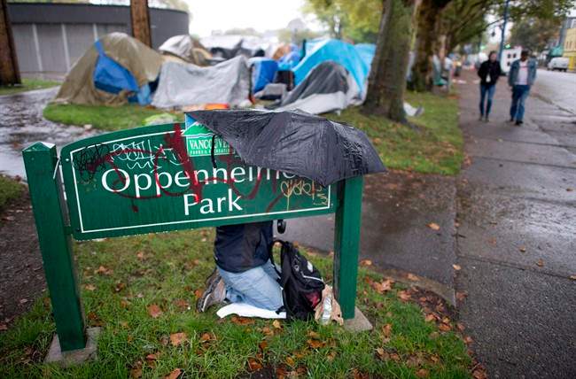 A woman takes cover from the rain behind a park sign in Oppenheimer Park in downtown Vancouver, Friday, Sept. 26, 2014. Vancouver's police force says some of its officers will be wearing video cameras during the dismantling of a homeless camp that is facing a court-ordered eviction. THE CANADIAN PRESS/Jonathan Hayward.