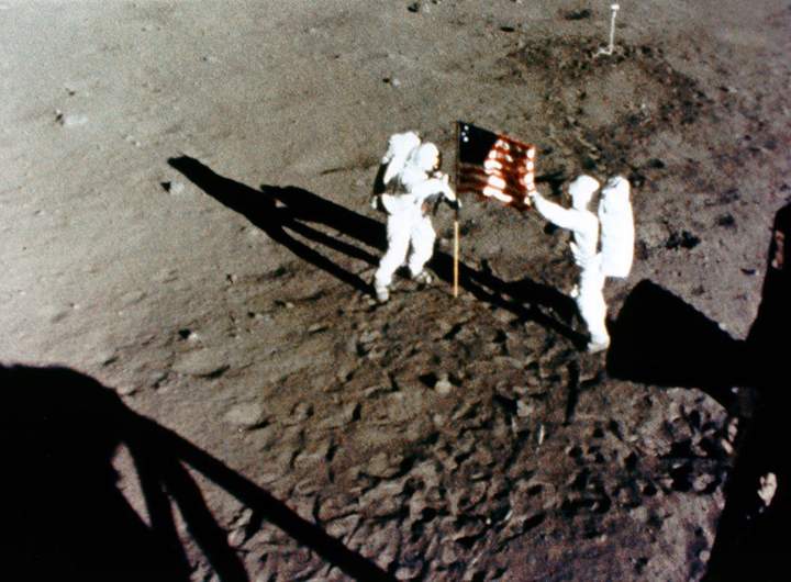 A camera mounted in the Lunar Module window photographs Neil Armstrong (left) and Buzz Aldrin (right) deploying the U.S. Flag on July 20, 1969.