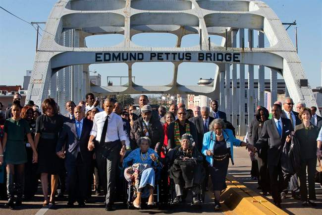 President Barack Obama, fourth from left, walks holding hands with Amelia Boynton Robinson, who was beaten during \”Bloody Sunday,\” as they and the first family and others including Rep. John Lewis, D-Ga, left of Obama, walk across the Edmund Pettus Bridge in Selma, Ala,. for the 50th anniversary of the landmark event of the civil rights movement, Saturday, March 7, 2015. At far left is Sasha Obama and at far right is former first lady Laura Bush. Adelaide Sanford also sits in a wheelchair.