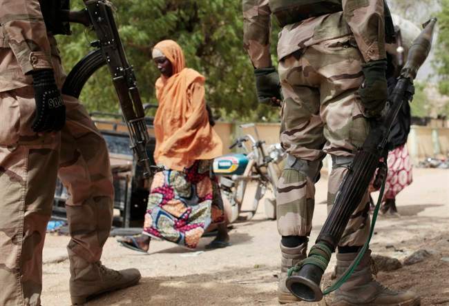 In this photo taken Wednesday, April 8, 2015, a woman walks past Nigerian Soldiers at a checkpoint in Gwoza, Nigeria, a town newly liberated from Boko Haram. 
