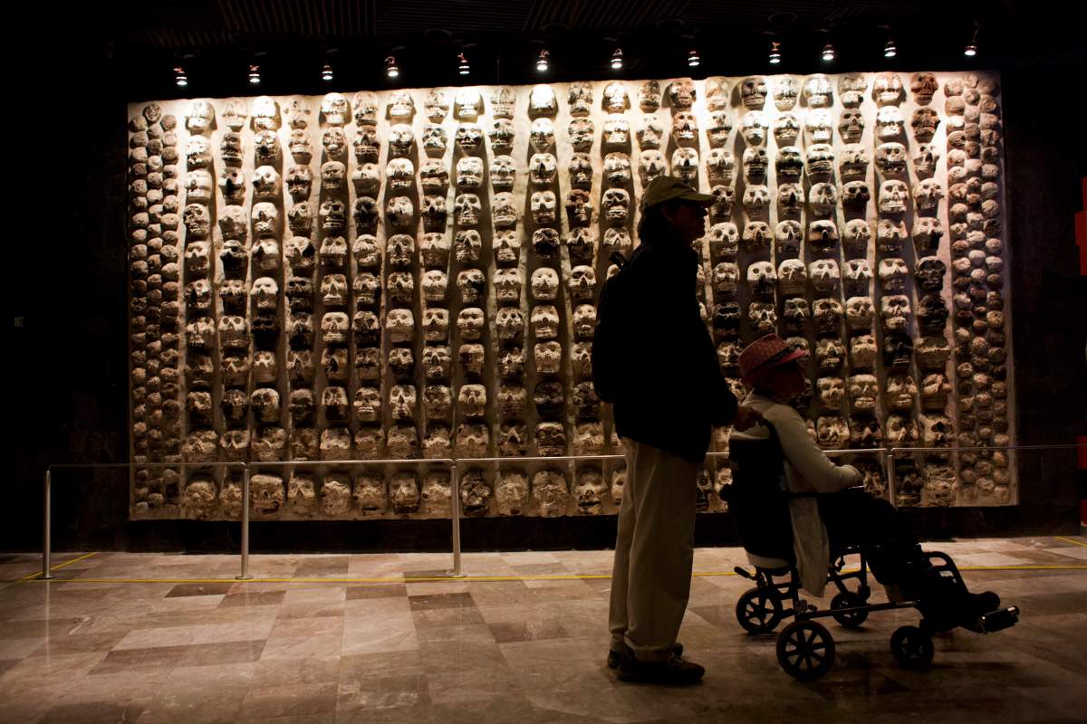 A man pushes a woman in a wheelchair past a wall of stone skulls that represent sacrificial victims, at the entrance to the Templo Mayor museum in central Mexico City, Friday, Aug. 7, 2015. The Templo Mayor museum has put on display for the first time an offering dedicated to Xochipilli, the Aztec god of singing, dancing, and the morning sun.