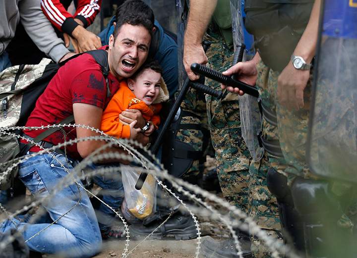 A migrant holding a child reacts as they are stuck between Macedonian riot police officers and migrants during a clash near the border train station of Idomeni, northern Greece on Friday, Aug. 21, 2015.