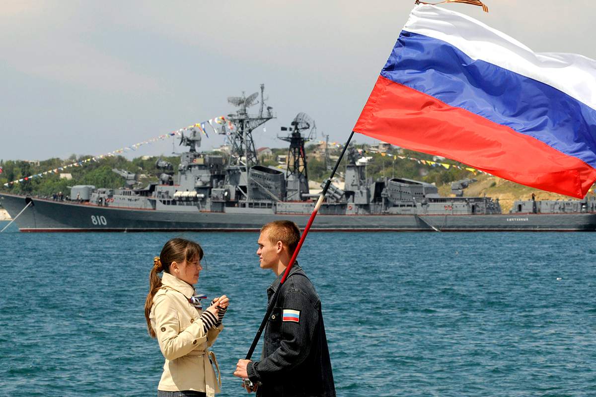 A man holding a Russian flag speaks with a girl wearing a telnyashka, a traditional navy sailor's striped vest, under her jacket during a naval parade to mark the 225th anniversary of Russian navy's Black Sea fleet at the Crimean Peninsula port of Sevastopol, Ukraine, Sunday, May 11, 2008, with the Russian Smetlivy destroyer in the background.  