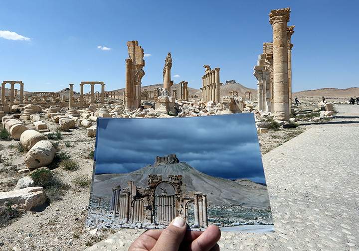 A general view taken on March 31, 2016 shows a photographer holding his picture of the Arc du Triomphe (Triumph’s Arch) taken on March 14, 2014, in front of the remains of the historic monument in the ancient Syrian city of Palmyra.