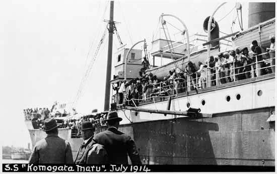Three men are pictured in front of the Komagata Maru, with passengers on deck. July 1914.