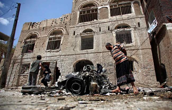 FILE - In this Tuesday, June 30, 2015 file photo, People stand amid wreckage of a vehicle at the site of a car bomb attack near a military hospital in Sanaa, Yemen.