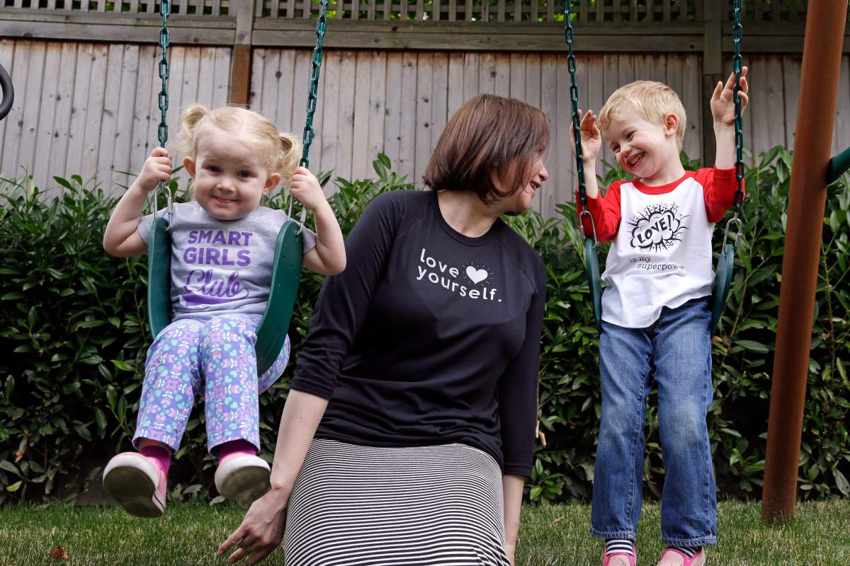 In this Thursday, July 7, 2016, photo, Courtney Hartman poses for a photo with her children Lois, 2, and Declan, 4, as they wear gender-neutral clothing of Hartman’s in Seattle. Hartman owns Jessy & Jack, a collection of unisex T-shirts, and Free to Be Kids, where a shirt with the slogan, ÄúI’m a Cat GuyÄù comes in blue, gray and yellow.