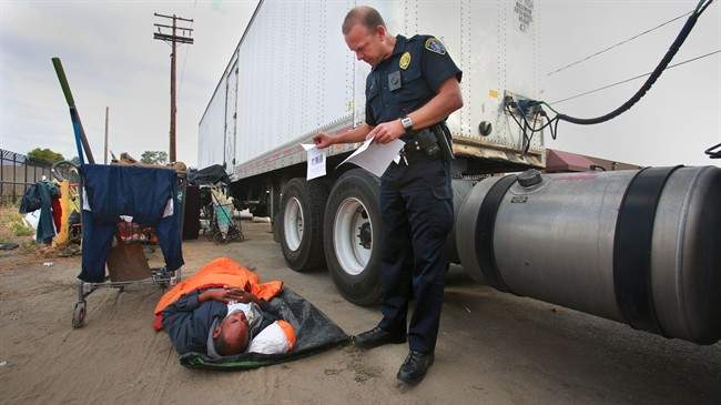 In this July 6, 2016, file photo, Ronald Mays, who rolls over to look at the flyer with pictures of a suspect in recent assaults on the homeless, as San Diego Homeless Outreach Team officer Brian Lucchesi canvasses several areas in the Midway and Sports Arena Blvd.