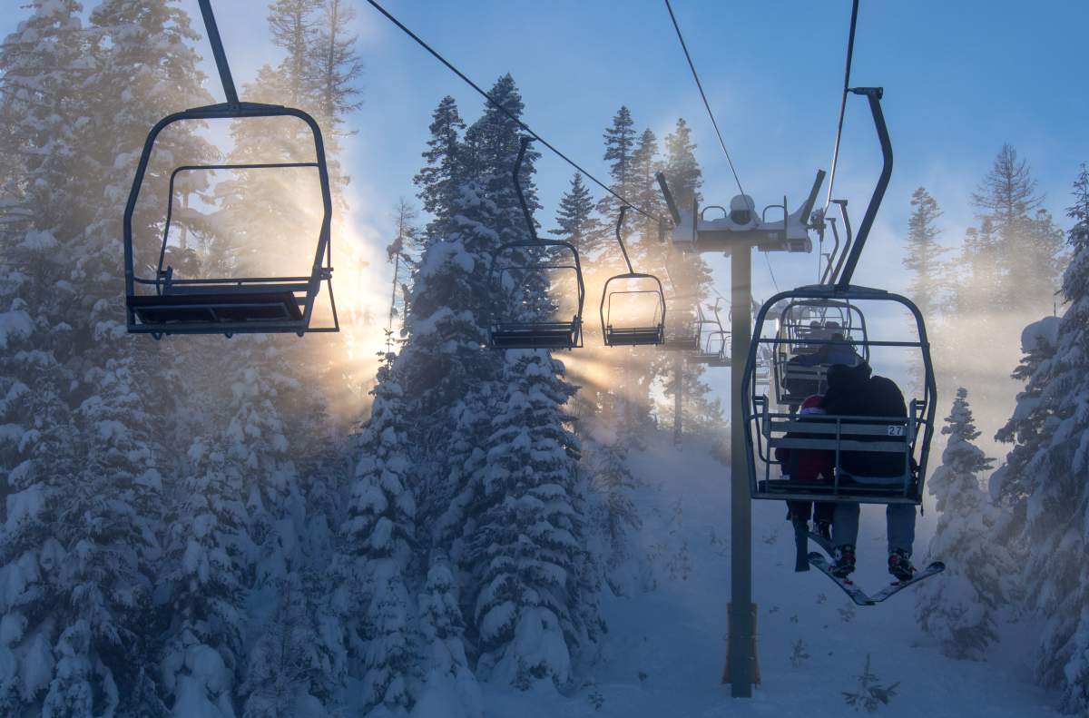 Sunbeams burst from behind a snow covered tree, viewed from a chairlift holding skiers.