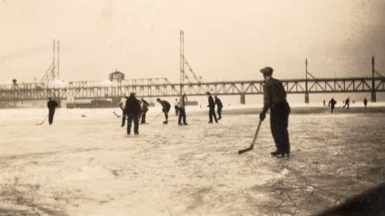 Photograph shows hockey being played on the frozen Fraser River by men and boys in the 1920s. The New Westminster Bridge can be seen in the distance.