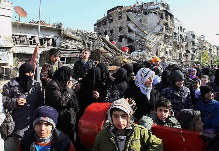 This Wednesday, Jan. 14, 2015 file photo, shows Syrians amid rubble as they wait in line at a military checkpoint in the town of Beit Sahm, south of the capital, Damascus. 