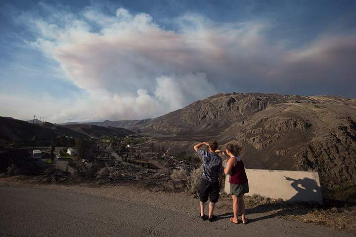 Ashcroft residents stop to view the remains of mobile homes destroyed by wildfire in Boston Flats, B.C., as a fire burns in the distance east of Cache Creek, on Sunday July 9, 2017.