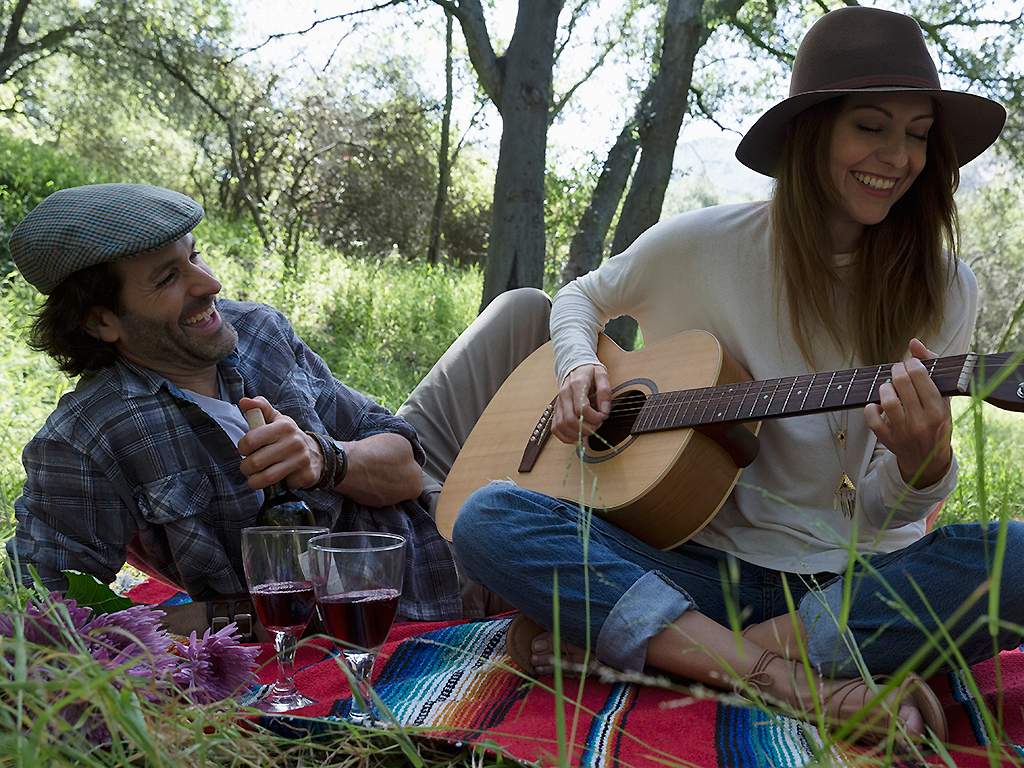 A couple drinks wine in the woods while playing guitar.