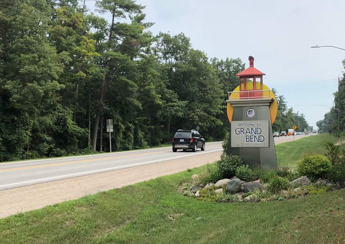 A tree-lined stretch of road with one vehicle on it passing a sign that looks like a lighthouse and says "Grand Bend" on it.
