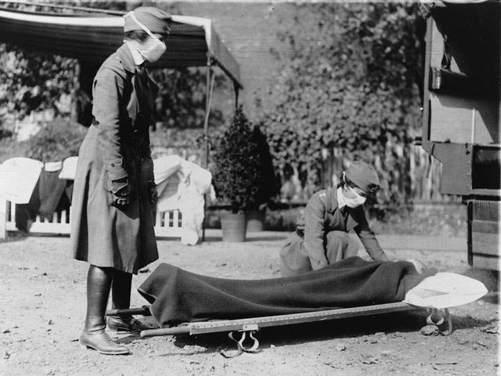 During the influenza epidemic, members of the Red Cross Motor Corps, both in uniform with cloth face masks, beside a stretcher on the ground, Washington DC, 1918.
