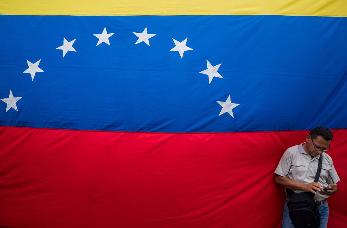 A man stands next to a Venezuelan flag as he looks at his cellphone during a signature collection event against the US blockade in Caracas, Venezuela, 15 August 2019. The President of Venezuela Nicolas Maduro suspended the talks with the opposition due to Juan Guaido's support of the blockade allegedely ordered by US President Donald Trump.