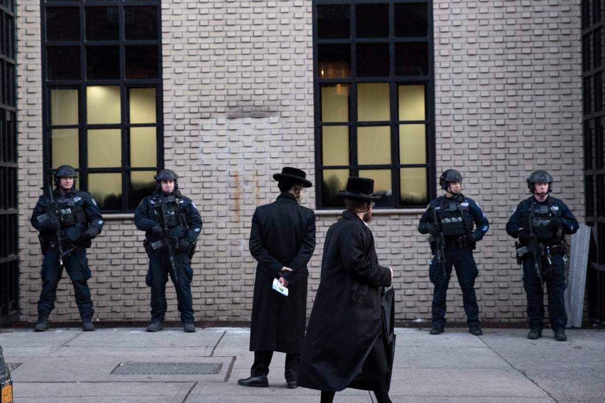 In this Dec. 11, 2019 photo, Orthodox Jewish men pass New York City police guarding a Brooklyn synagogue prior to a funeral for Mosche Deutsch in New York.