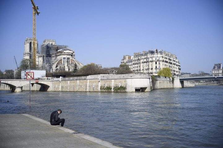 A man sits on the banks of the River Seine near the ruins of Notre Dame Cathedral in Paris on March 19, 2020.