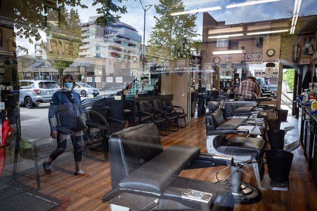 A man cleans inside a closed barbershop as a pedestrian wearing a face mask is reflected in the shop's window, in Burnaby, B.C., on Wednesday, May 6, 2020. Salons and barbershops are starting to reopen in some parts of Canada with more on the way, but the industry says that the measures needed to keep everyone safe while getting a haircut will mean a big financial hit. THE CANADIAN PRESS/Darryl Dyck.