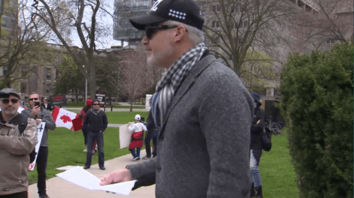 A man speaks to protesters at Queen’s Park as the Canadian flag is held upside down in the background.