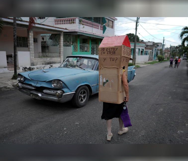 Retired nurse Feridia Rojas, 82, walks past a vintage car as she wear cardboard, shaped into a house, to protect herself from COVID-19. The box reads: “I am at home. And you?”