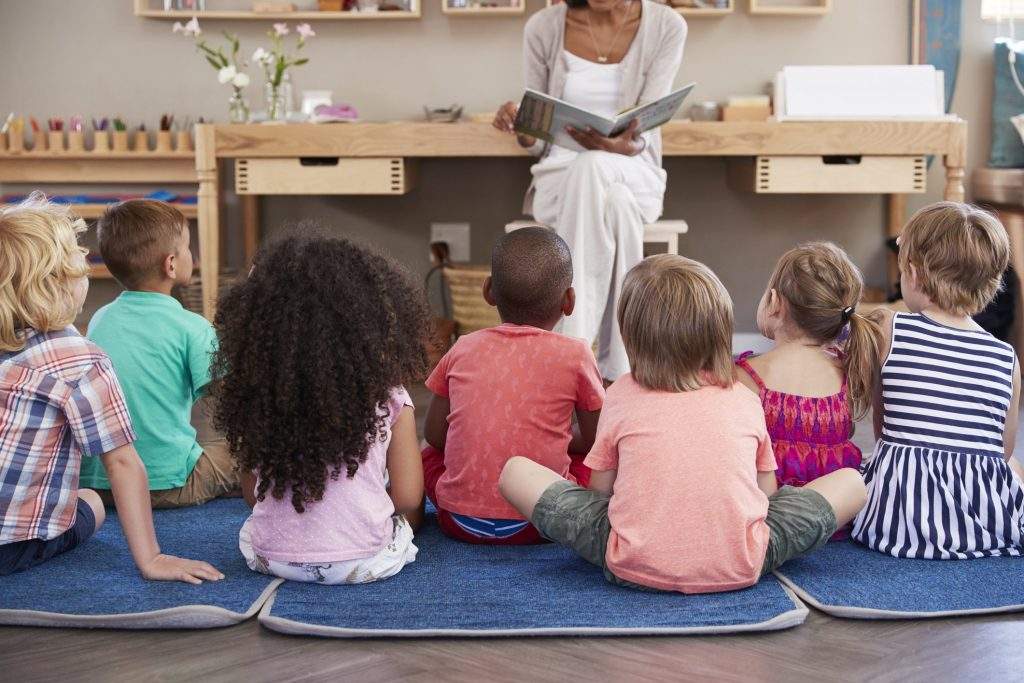 Teacher At Montessori School Reading To Children At Story Time.