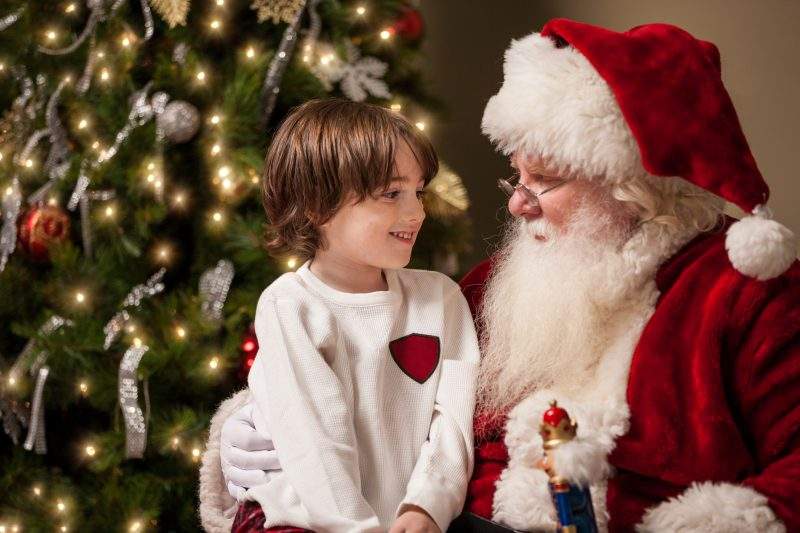 A young boy sits on Santa Claus' lap by a decorated Christmas tree. 