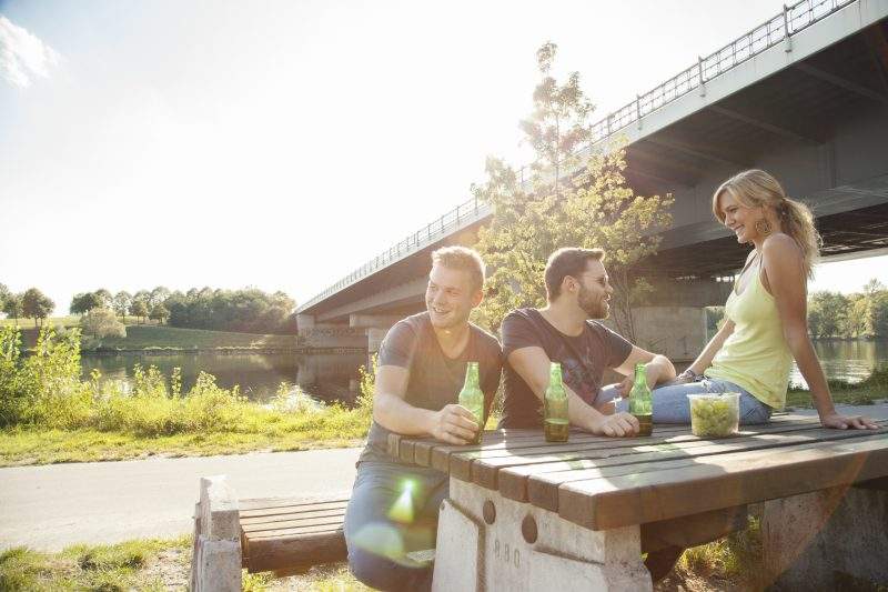 Three young friends drinking beer on riverside picnic table.