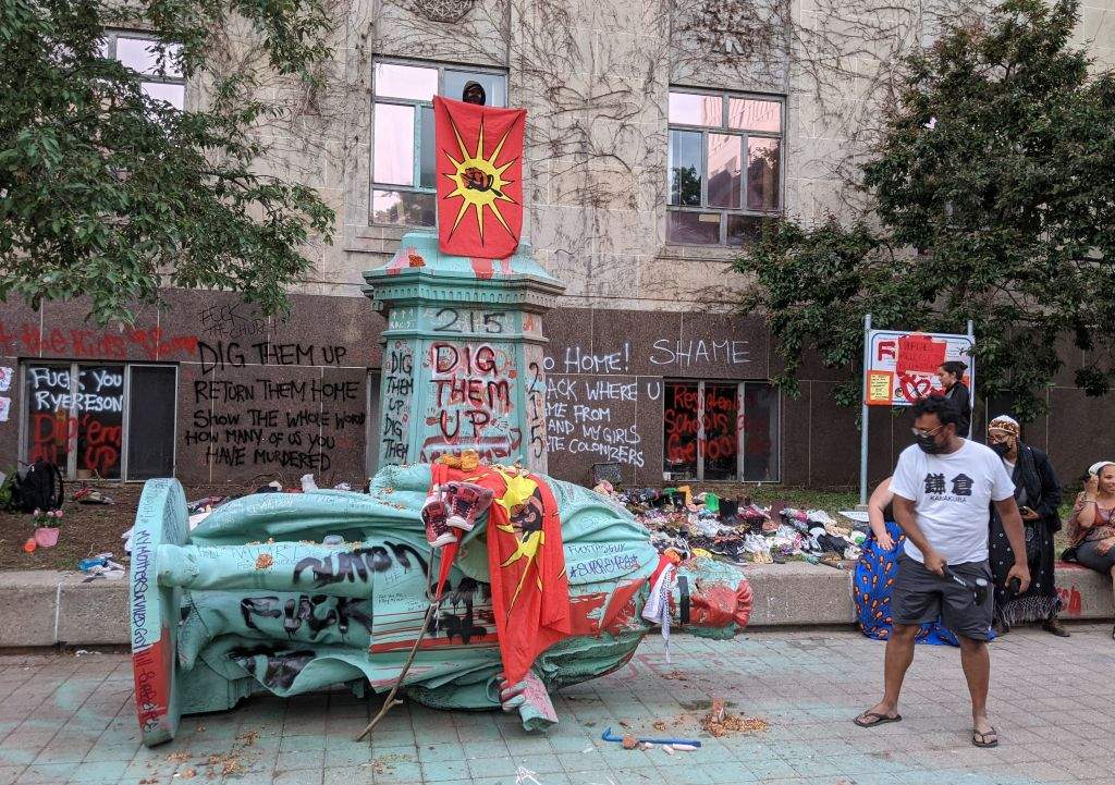A man waving the Mohawk Warrior Society flag stands on the pedestal of the toppled statue of Egerton Ryerson, while another man hits it with a hammer in Toronto on June 6, 2021.
