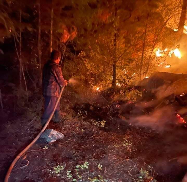 A homeowner douses hot spots with a hose as the Nk\’Mip Creek wildfire burns within 100 feet of his property on Wednesday night, July 21, 2021.