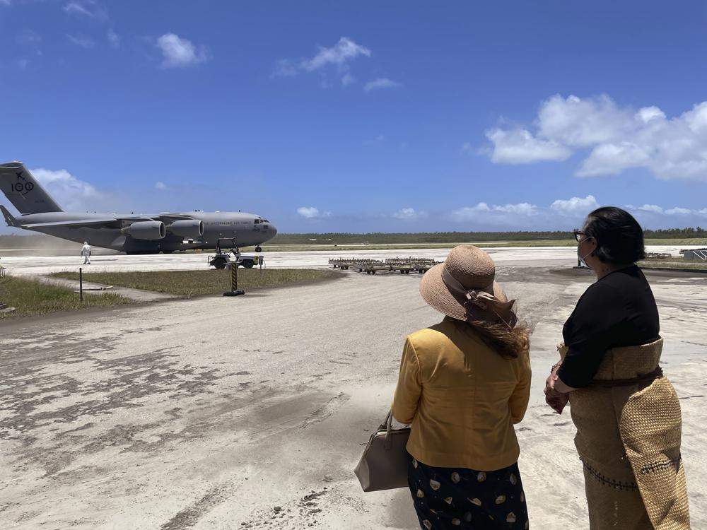 Tongan officials stand on a runway, watching a plane arrive with aid supplies.