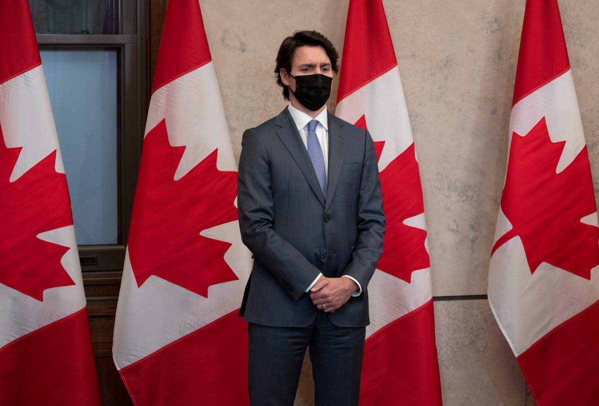 Prime Minister Justin Trudeau stands in front of a row of red and white Canadian flags. He stares off ahead, wearing a black face mask and a dark suit, with his hands clasped in front of him.