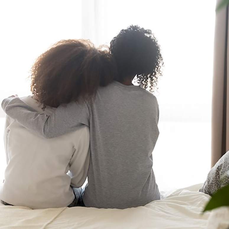 two Black women with their backs to the camera, sitting on a bed and comforting one another