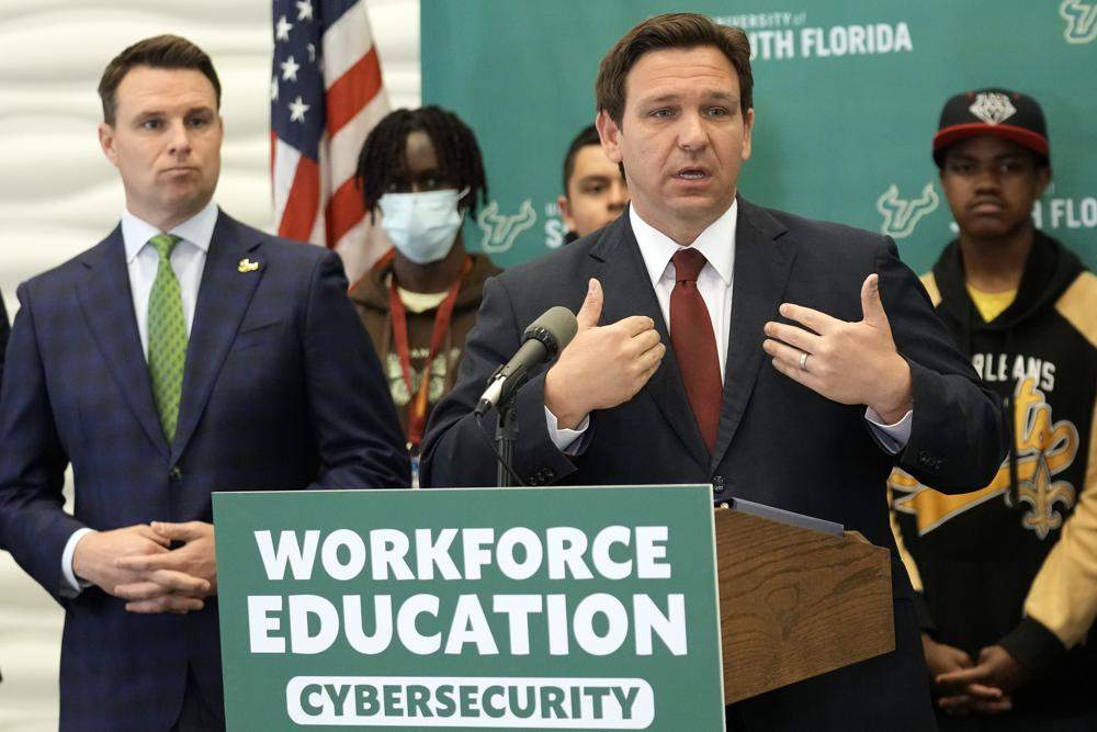 Ron DeSantis stands behind a lectern wearing a suit and tie at a press conference. Students stand behind him.
