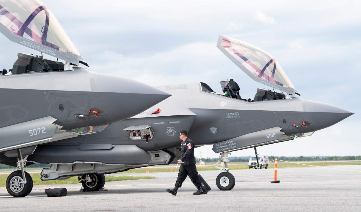 The F-35 fighter jet is seen while on the tarmac at an Ottawa airport. It is slate grey against a cloudy sky, with one person walking by the wheels.