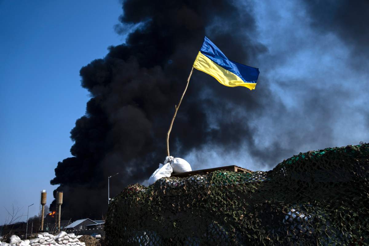 A blue and yellow Ukrainian flag flies alone against a plume of black, billowing smoke caused by a Russian attack.