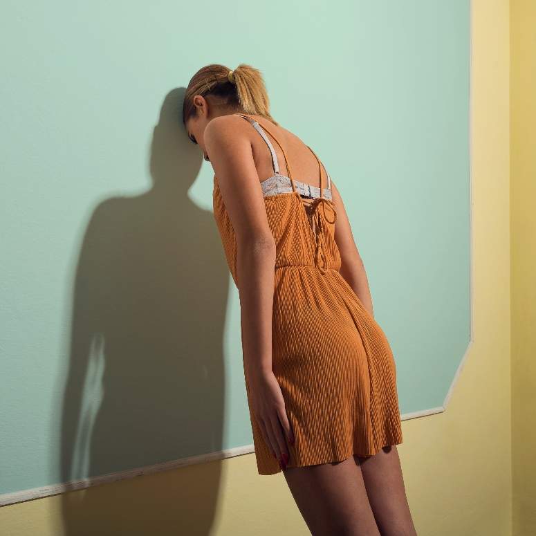 A woman in an orange romper leaning with her forehead against a light blue and yellow wall