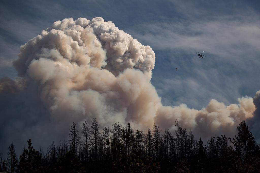 A helicopter carrying a water bucket flies past a pyrocumulus cloud, also known as a fire cloud, produced by the Lytton Creek wildfire burning in the mountains above Lytton, B.C., on Sunday, August 15, 2021. The Insurance Bureau of Canada says there is now a path to begin the recovery of the Village of Lytton, almost a year after two people died and most of the community was destroyed by a wildfire. THE CANADIAN PRESS/Darryl Dyck.