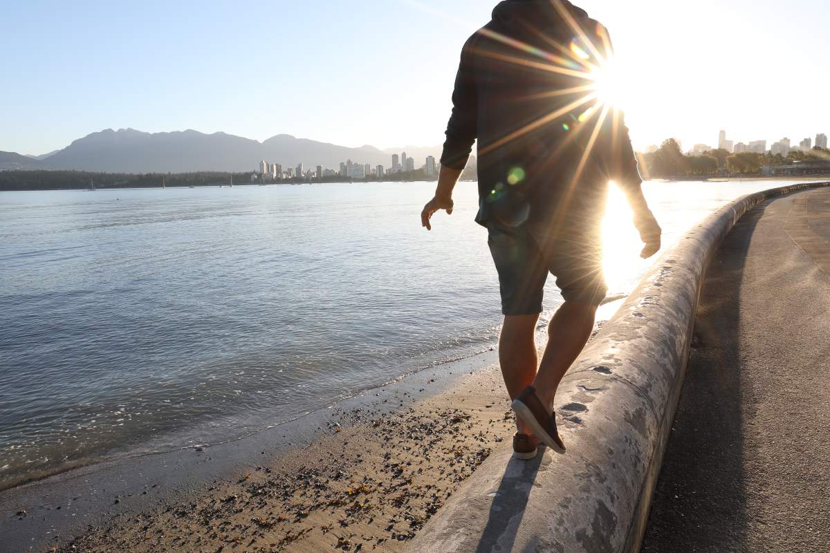 Man balances on stone wall in waterfront area, sun in front of him.