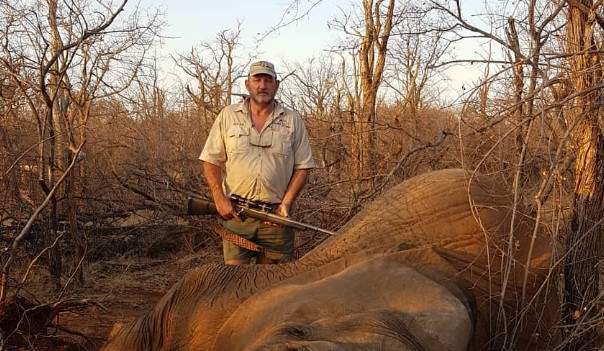 Riaan Naude poses alongside a dead elephant. He is wearing a white shirt and holding a gun.