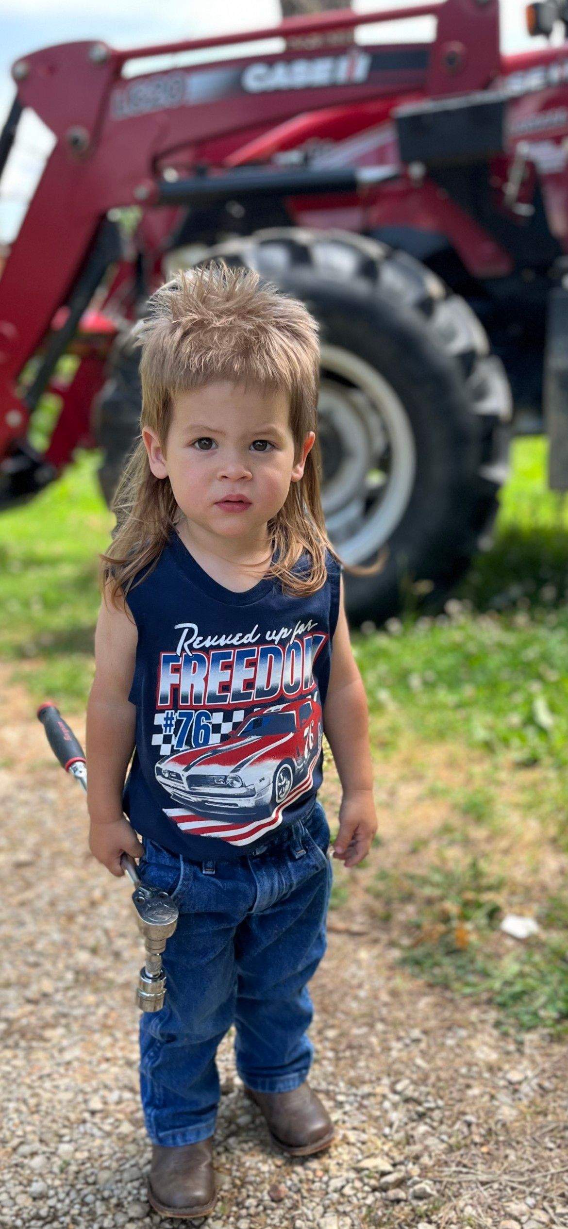 A young child with a mullet poses in front a a car.