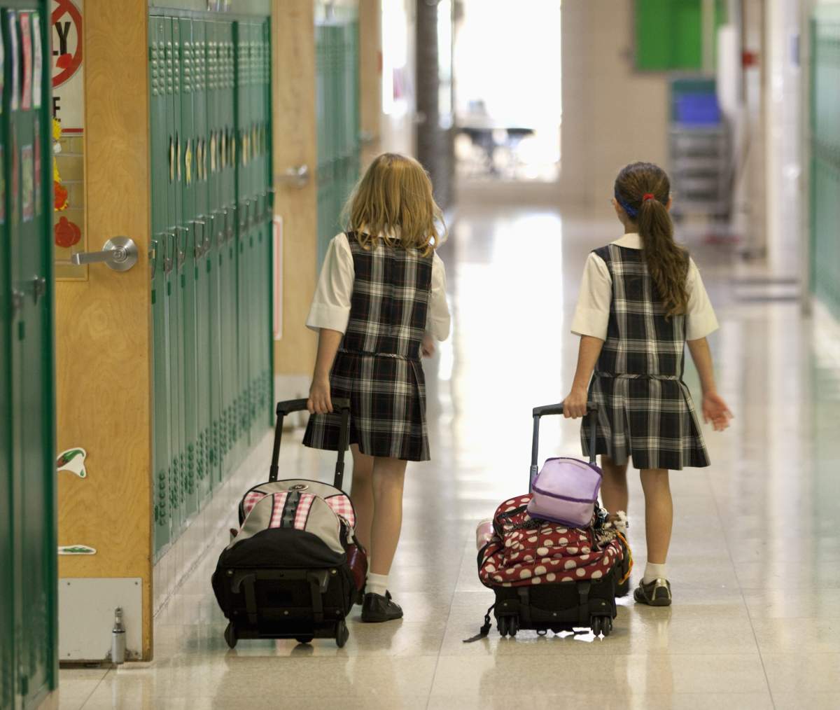 Eight year old girls head home after after school rolling their backpacks down the school hallway.
