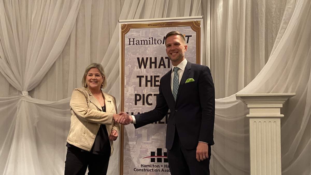 Andrea Horwath and Keanin Loomis stand in front of a sign for HamiltoNEXT that says "WHAT'S THE BIG PICTURE HAMILTON?" while smiling and shaking hands.