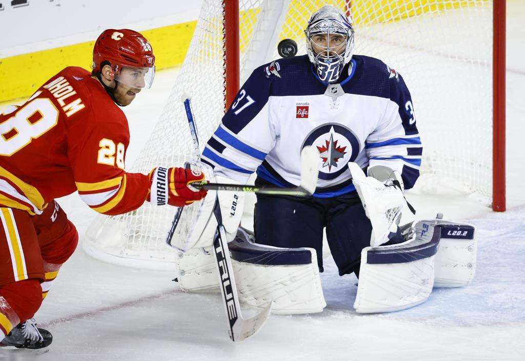 Winnipeg Jets goalie Connor Hellebuyck, right, follows the puck as Calgary Flames forward Elias Lindholm tries to get his stick on it during third-period NHL pre-season hockey action in Calgary, Friday, Oct. 7, 2022.
