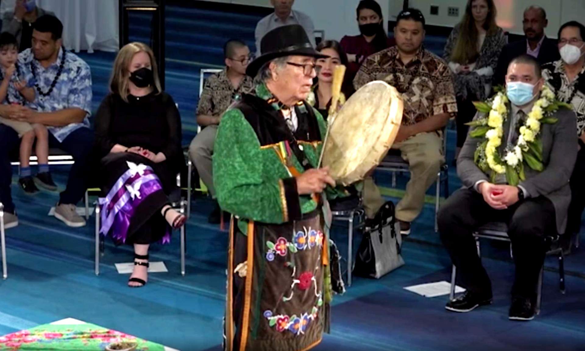An Indigenous drumming ceremony is performed by a solo drummer who is standing in the centre of a circle. The audience is seated, looking on.