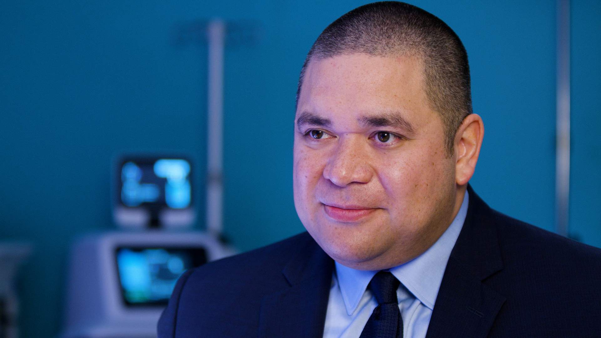 Dr. Alika Lafontaine has closely shaved hair and is wearing a dark suit and tie with a light coloured dress shirt. He is looking towards the left of the camera lens during an interview in a dimly lit operating room.